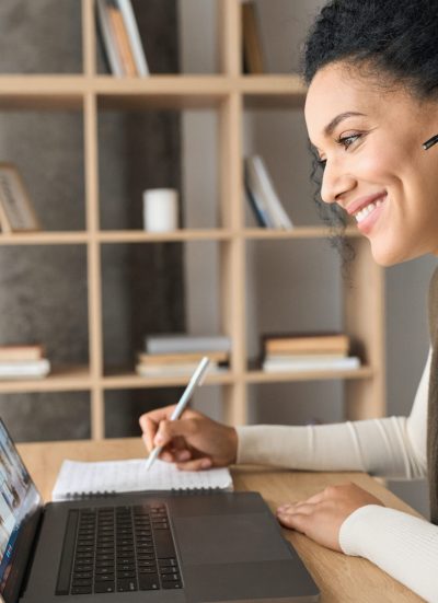 African American female student having video call videoconference on laptop. African American mixed race adult student wearing headset having virtual meeting online call educational webinar chatting at home office writing notes. Video e learning conference call on pc.