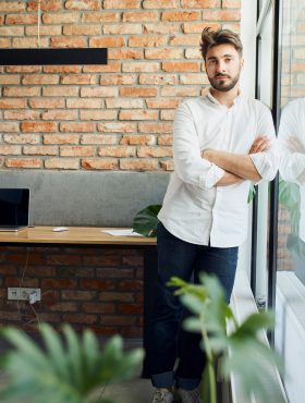 Young man entrepreneur looking at camera standing in small startup office Young man entrepreneur looking at camera standing in small startup office